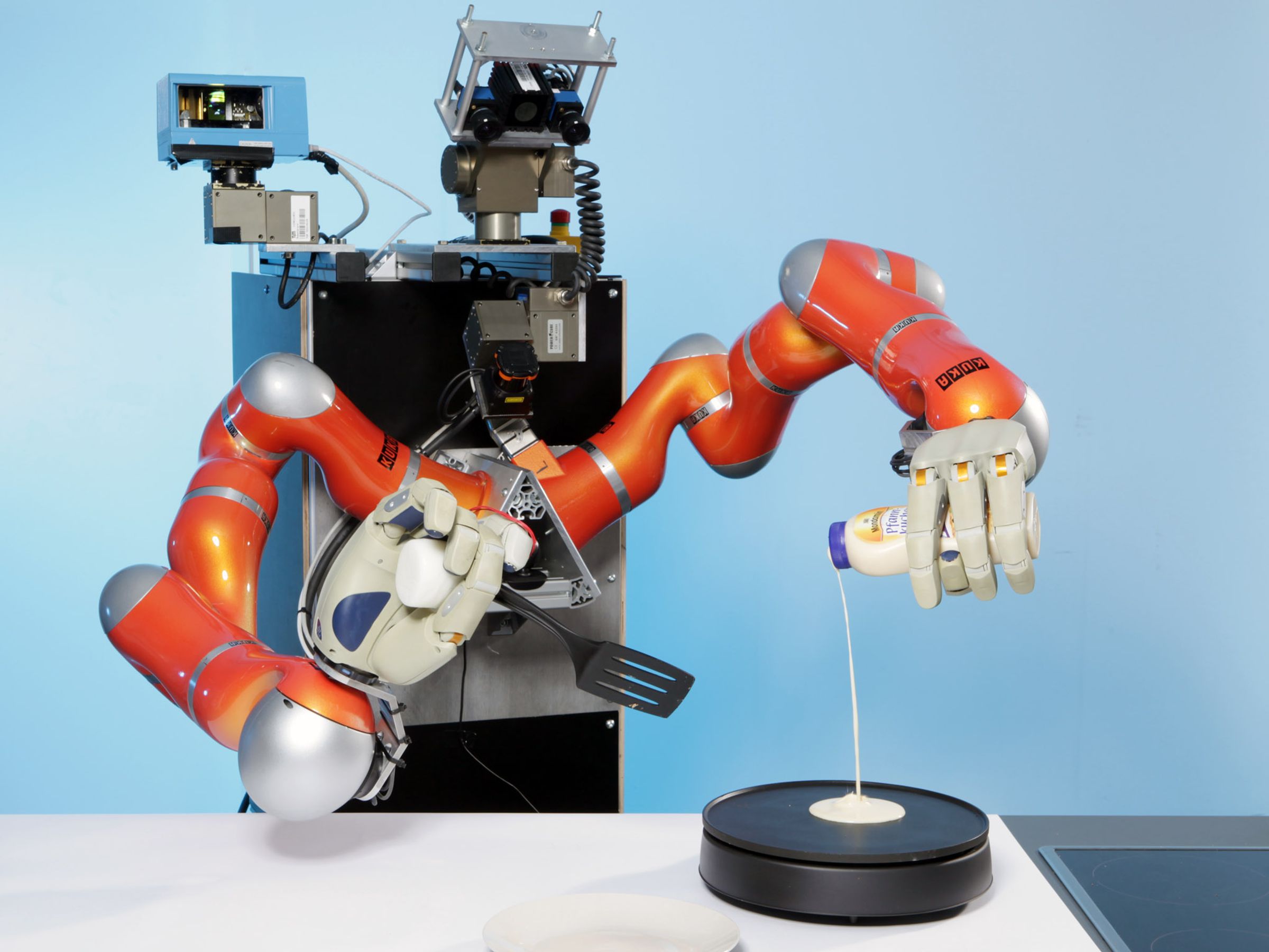 A tall humanoid robot with two orange arms and large robotic hands holds a spatula and pours batter on a hot plate to make pancakes or crepes in a kitchen.
